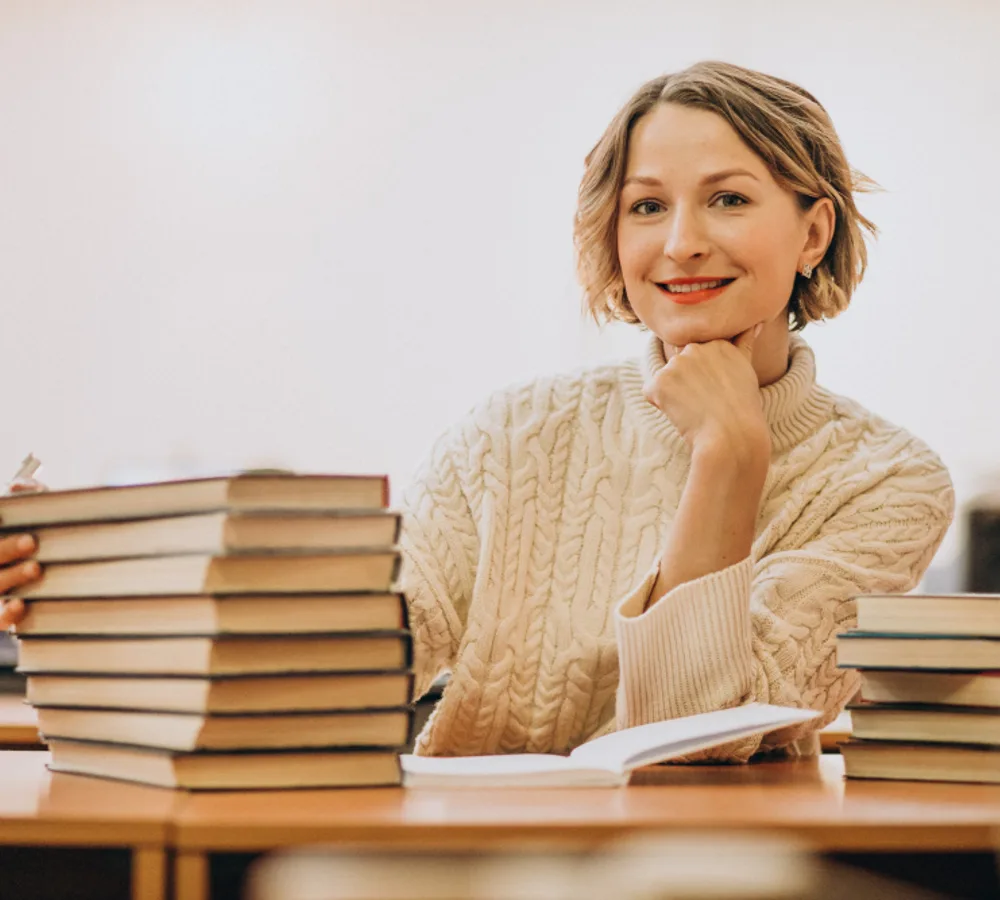 Uśmiechnięta nauczycielka siedząca przy stole ze stosami książek w bibliotece